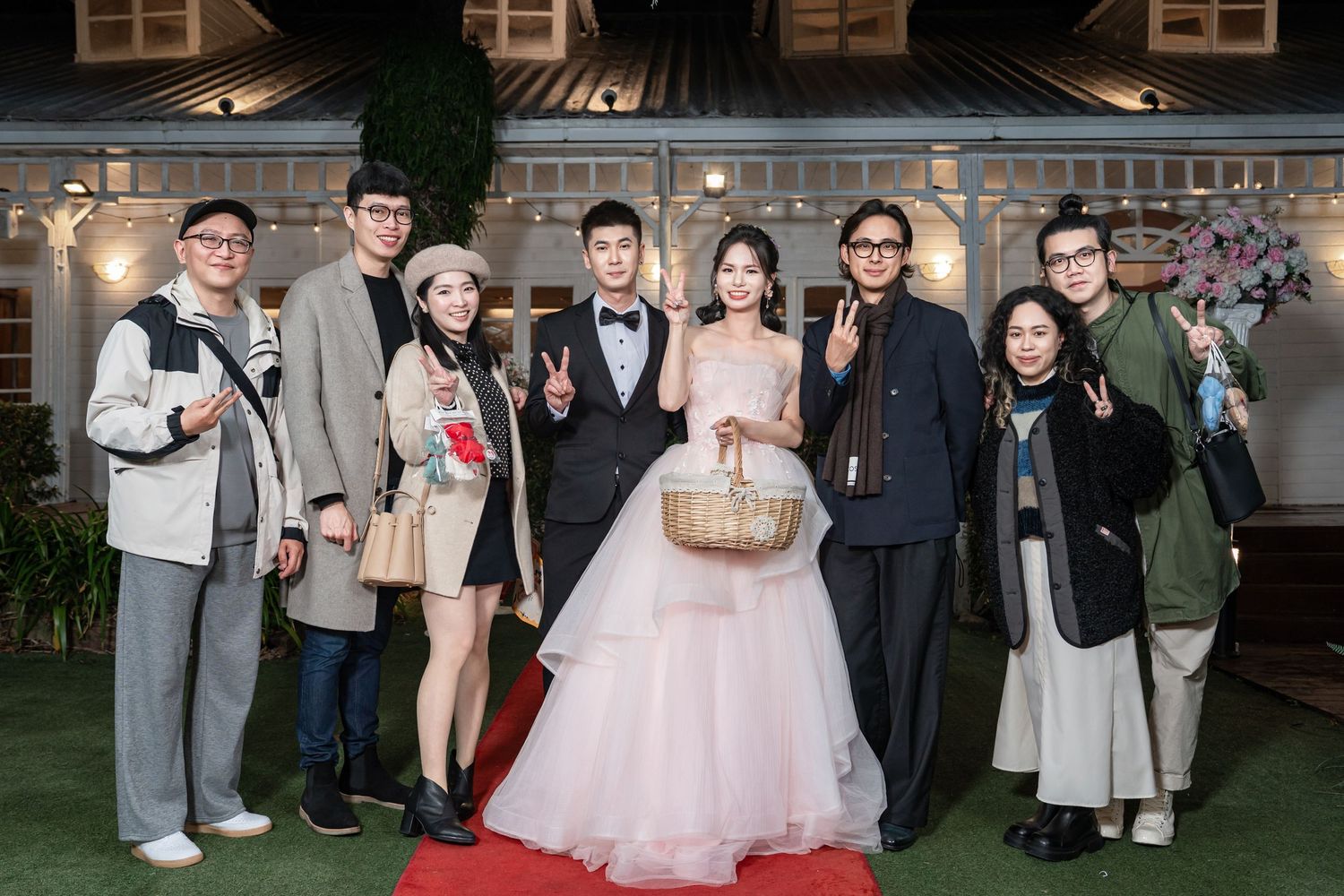 A group of people pose together at a formal event, with a person in a pink wedding gown holding a basket in the center.
