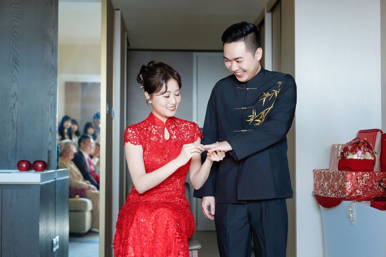 A couple in traditional Chinese wedding attire, with the bride in red qipao and groom in black suit examining something together.