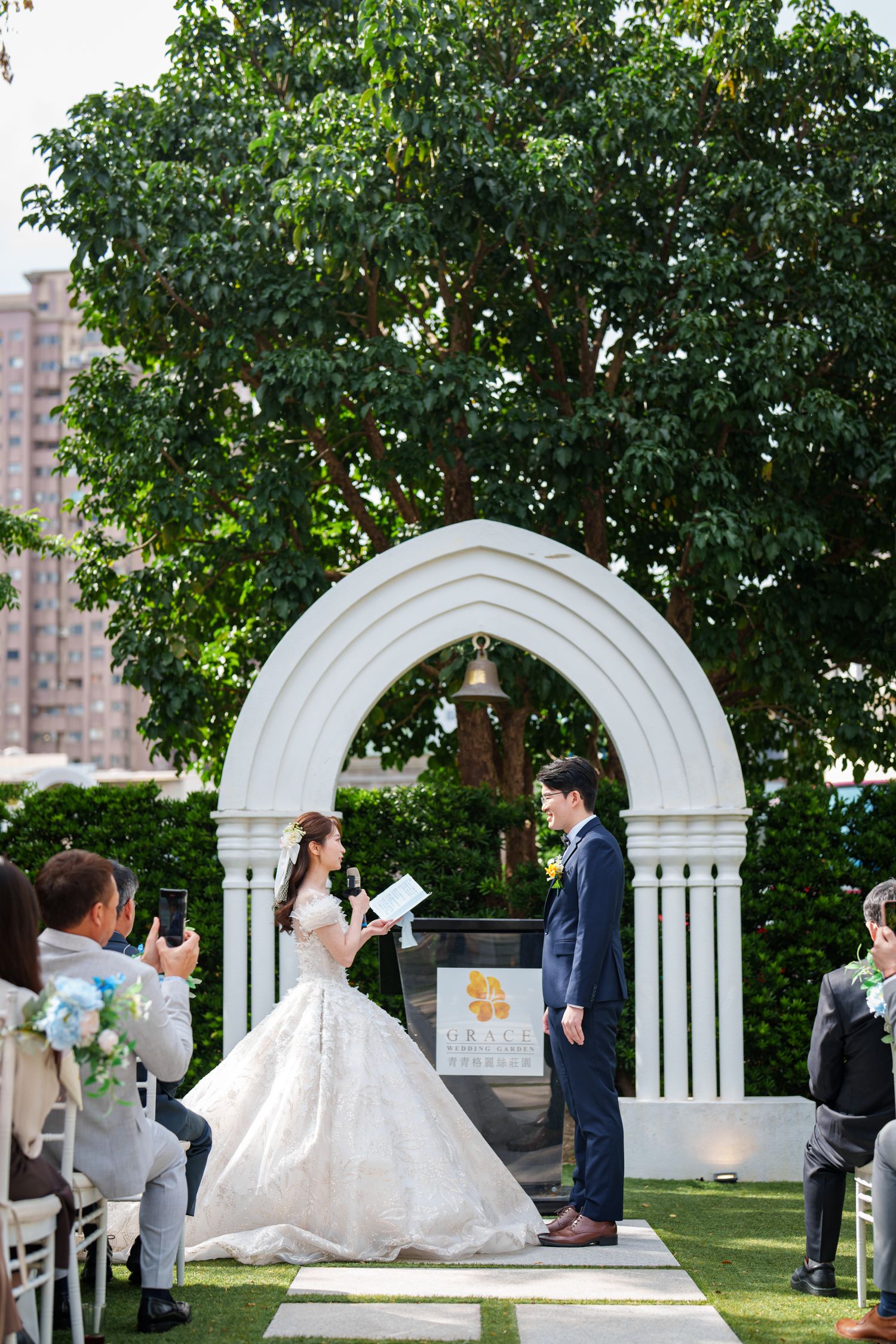 A wedding ceremony takes place under a white garden arch surrounded by lush greenery and seated guests.