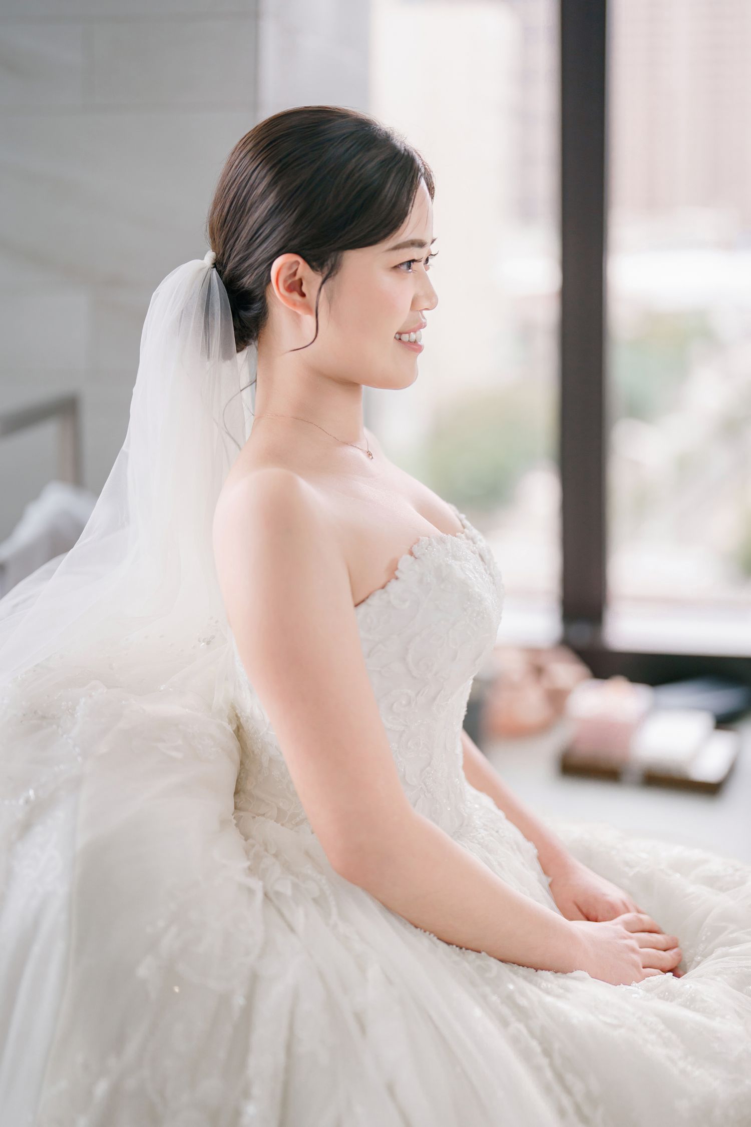 A bride in a white wedding dress poses gracefully by a window in soft natural light.