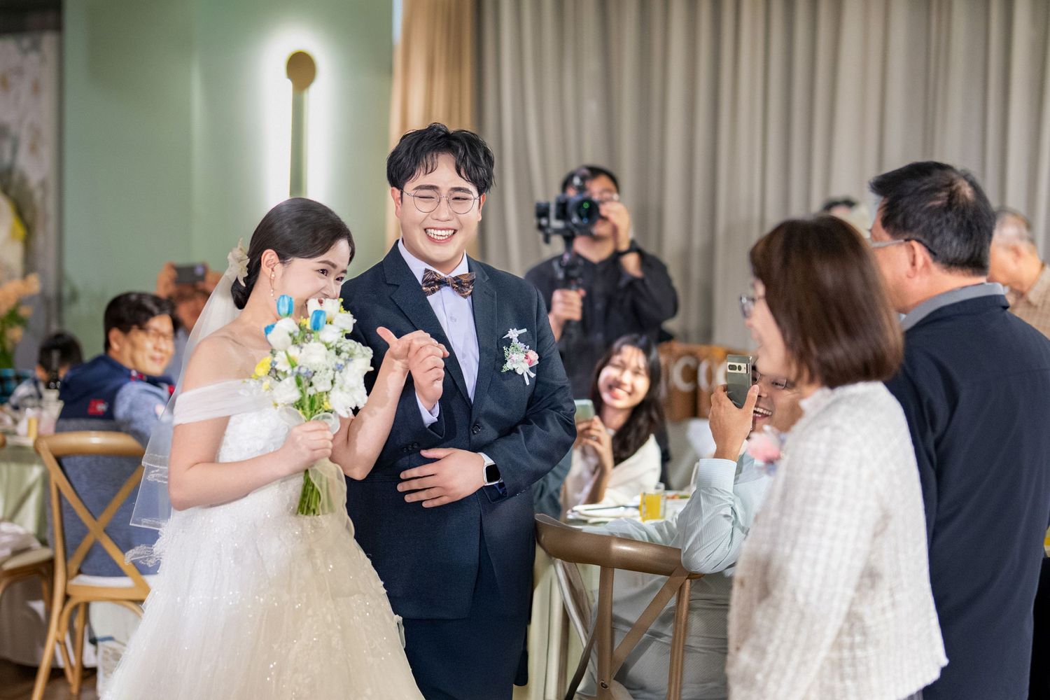 Wedding reception guests enjoy dancing and celebrating at an elegant indoor venue with white curtains and decor.