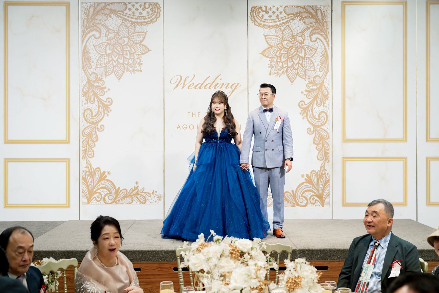 Wedding reception in an elegant white and gold ballroom with guests seated at round tables and blue decor accents.