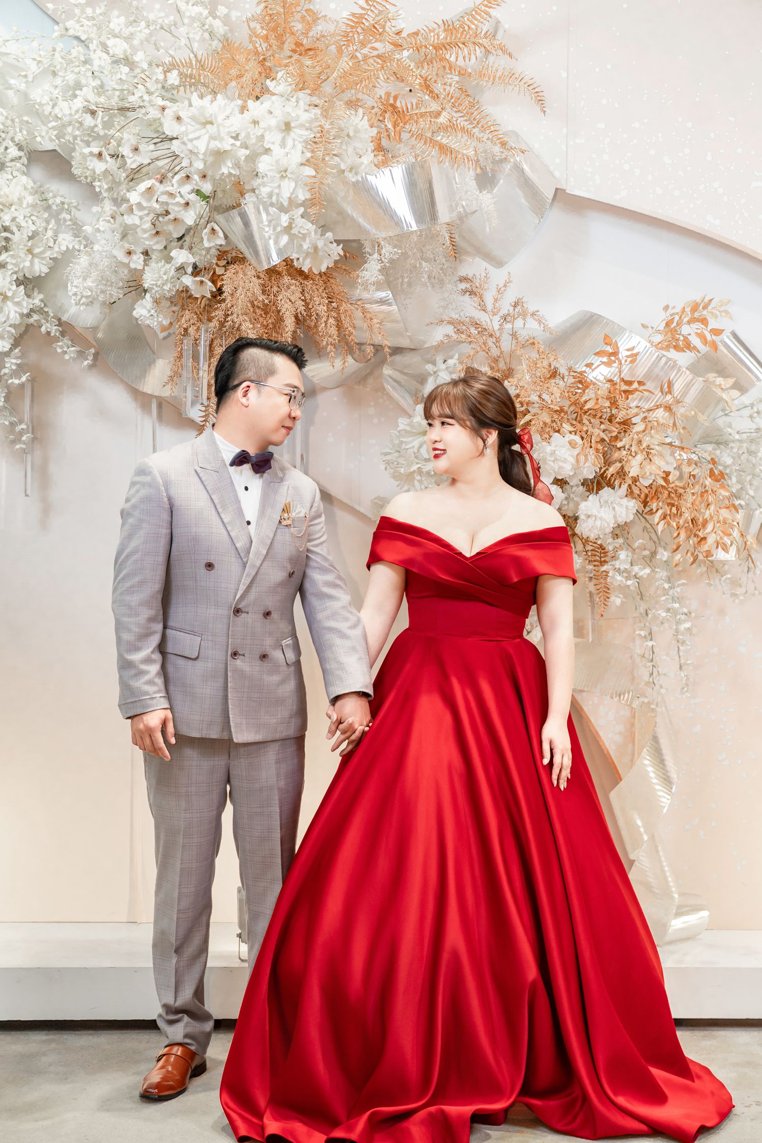 A couple in a gray suit and red ballgown share joyful moments against a white floral backdrop during their wedding photoshoot.