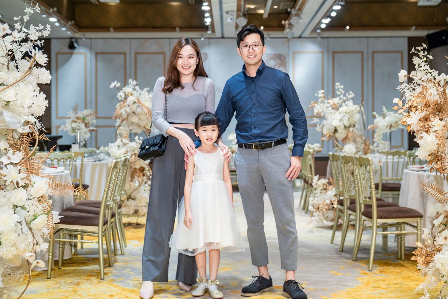 Family poses together in an elegant banquet hall with floral decorations and gold chairs in the background.