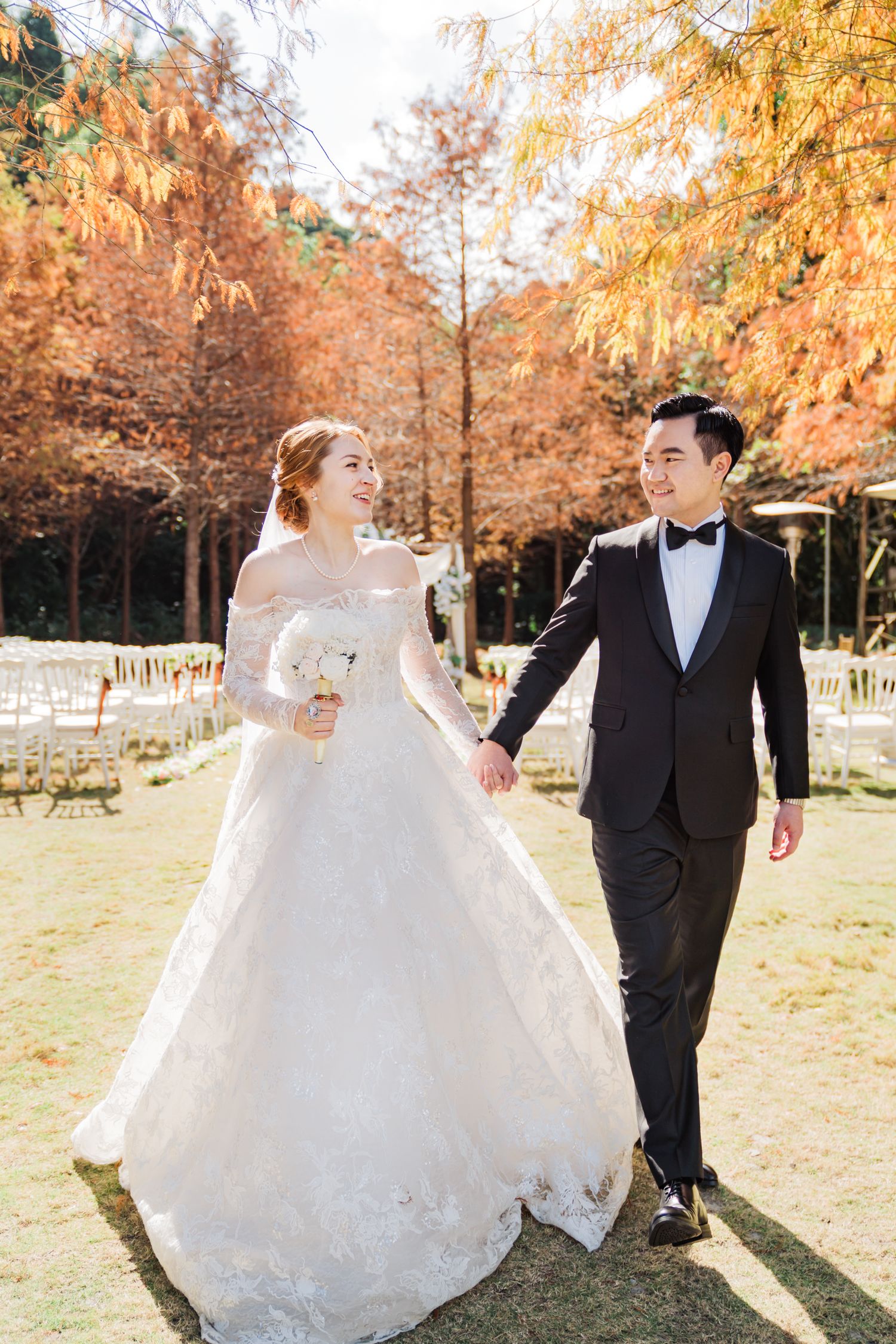 Elegant outdoor autumn wedding scene with white lace gown and black tuxedo among golden trees.