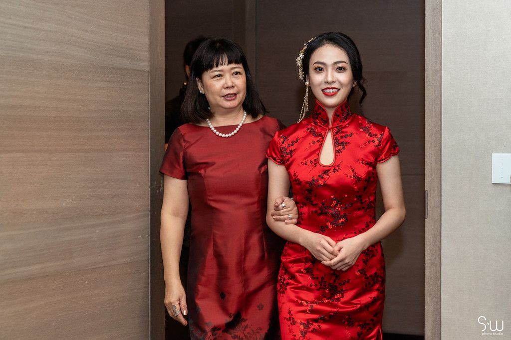 Two women wearing traditional red Chinese qipao dresses stand together in an interior hallway.