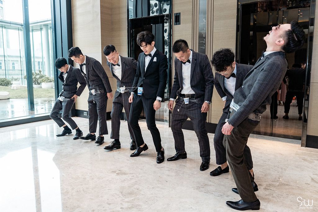 Wedding party members in suits perform a synchronized leaning pose in a hallway.