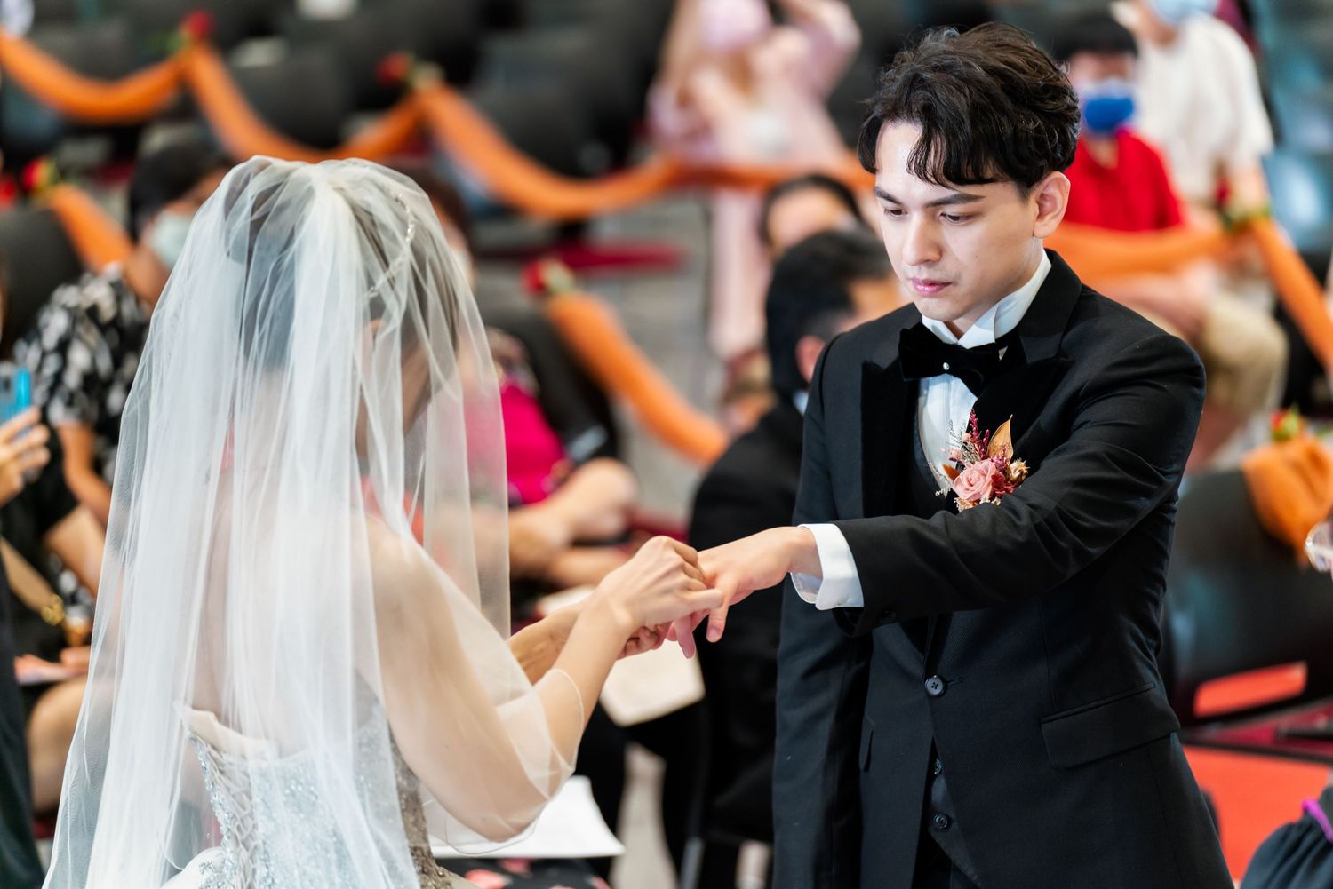 A bride and groom exchange rings during their wedding ceremony in an auditorium.