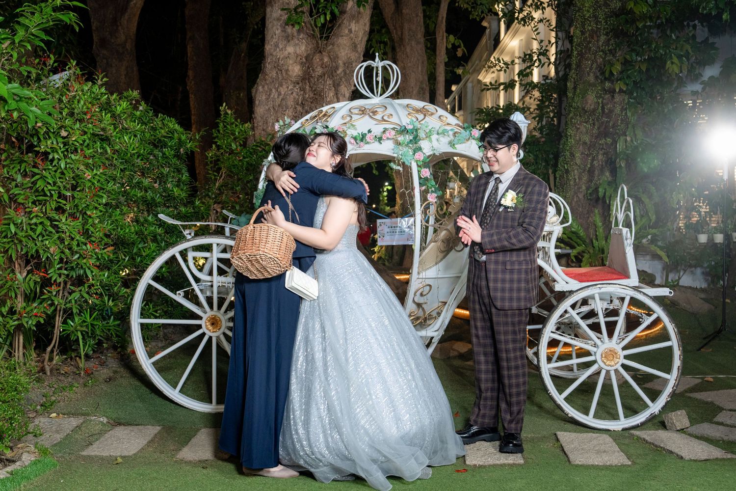 A wedding party poses in a garden setting with a decorative white carriage wheel visible in the background.
