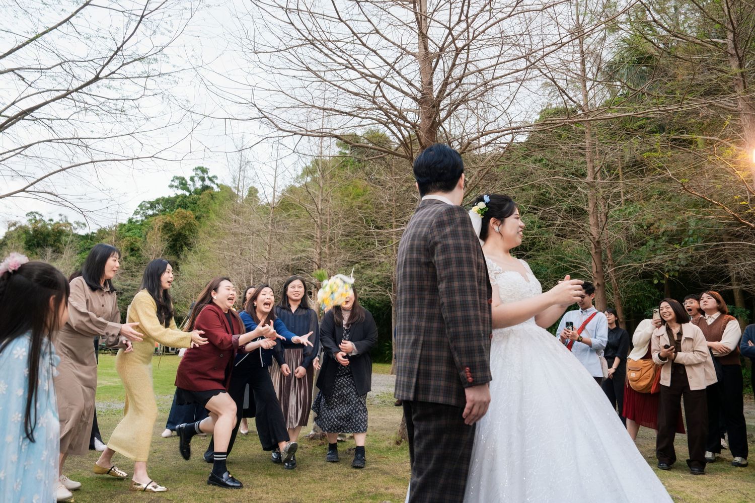 A group of wedding guests gather outdoors in a wooded area during a spring celebration.