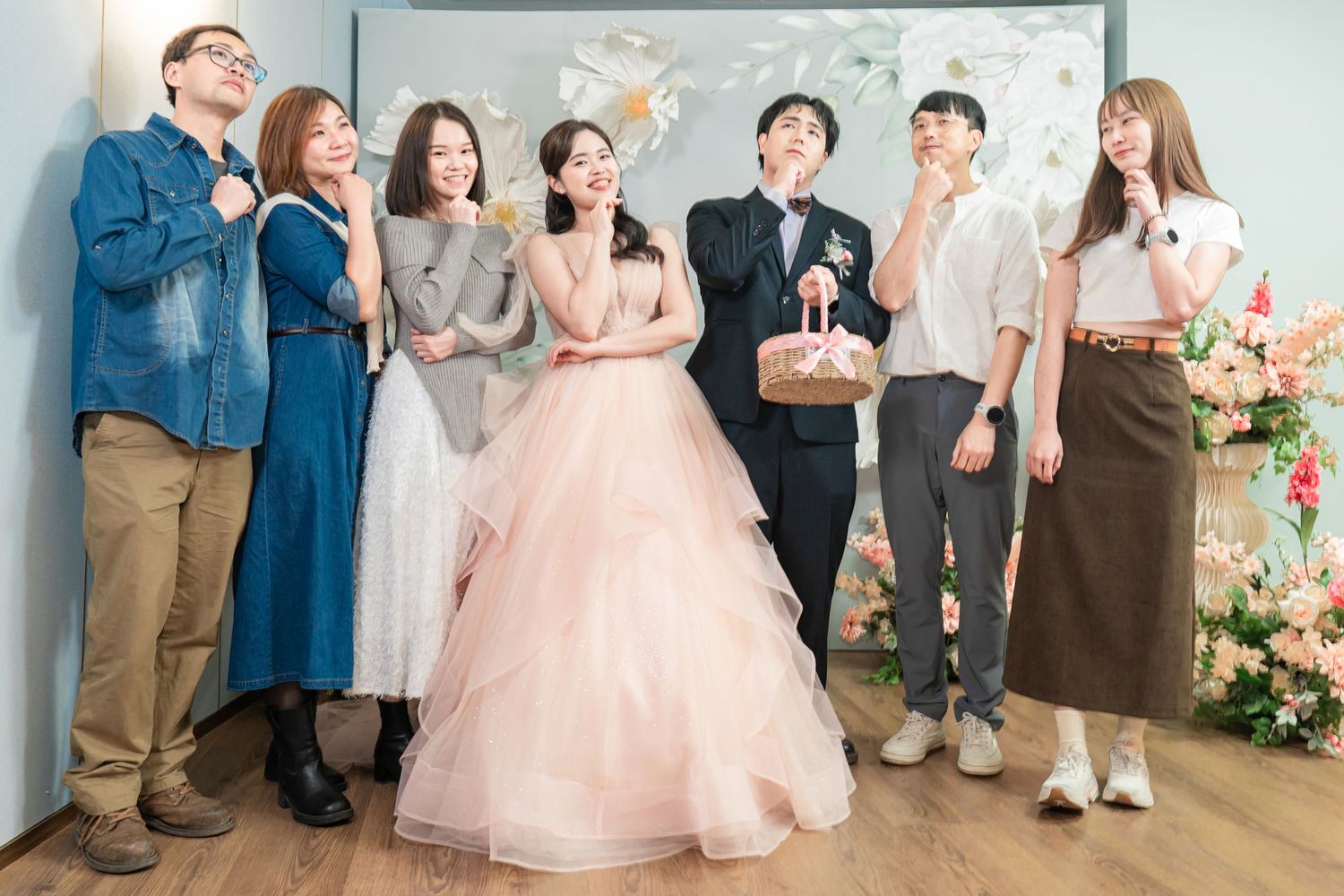 Wedding party members in formal attire stand in line against a white wall with floral decorations.
