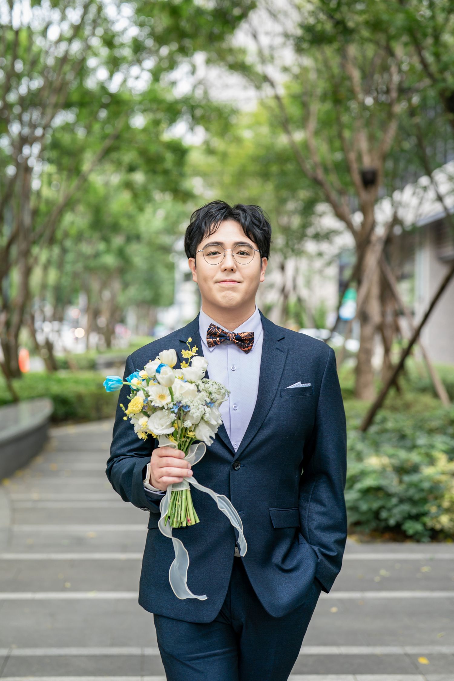 A person in a formal black suit holds a champagne glass and wedding bouquet in an outdoor garden setting.