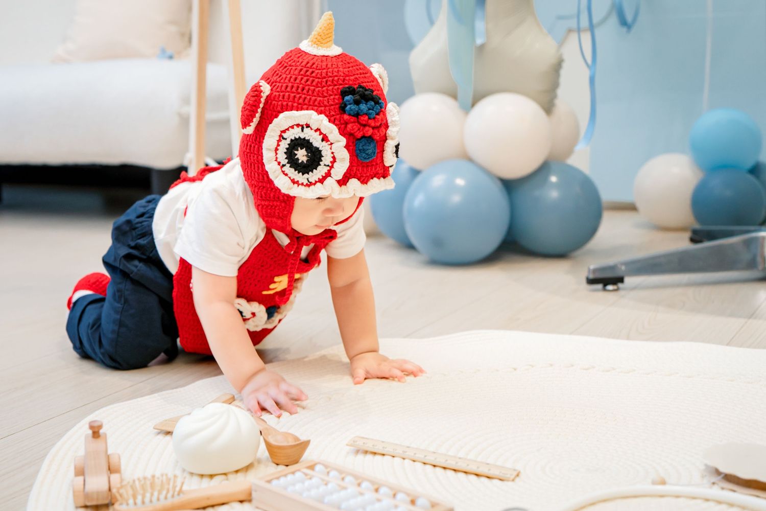 Baby in a red crochet owl hat crawls on white floor surrounded by blue and white balloons during a photo session.