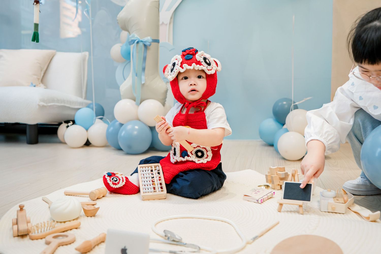 Small child in a red and white outfit plays with toys on a white floor surrounded by soft blue decor.