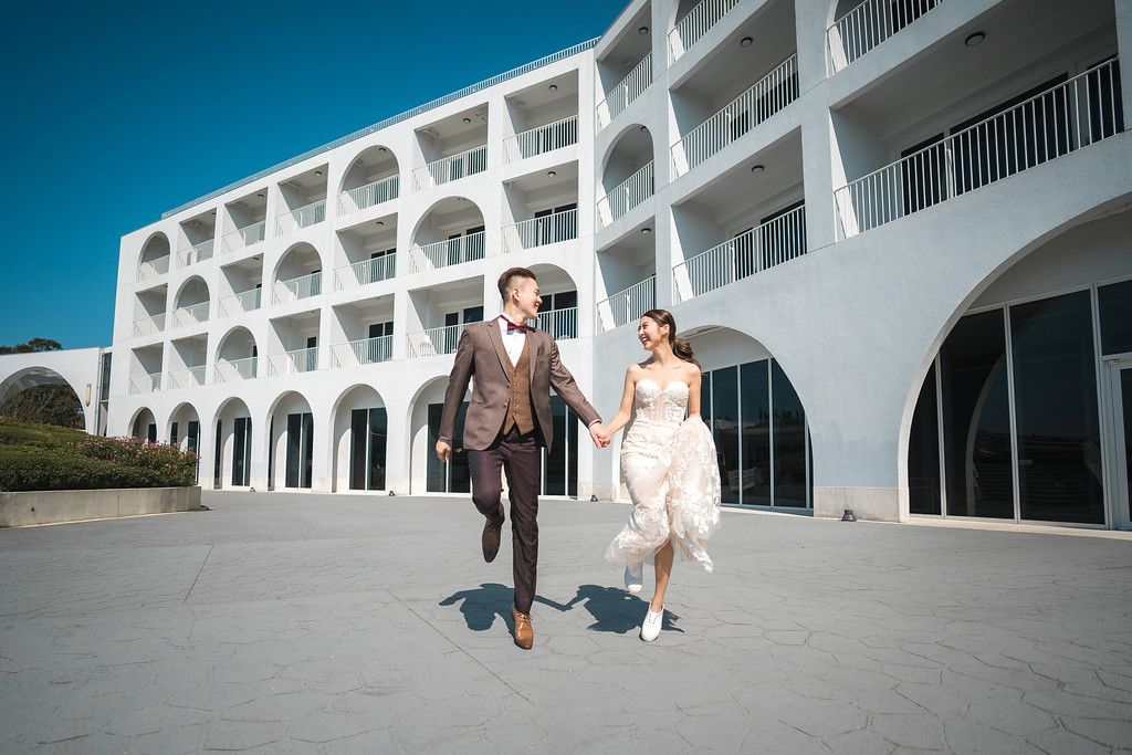 Couple walking along a modern white architectural corridor with arched windows and columns.
