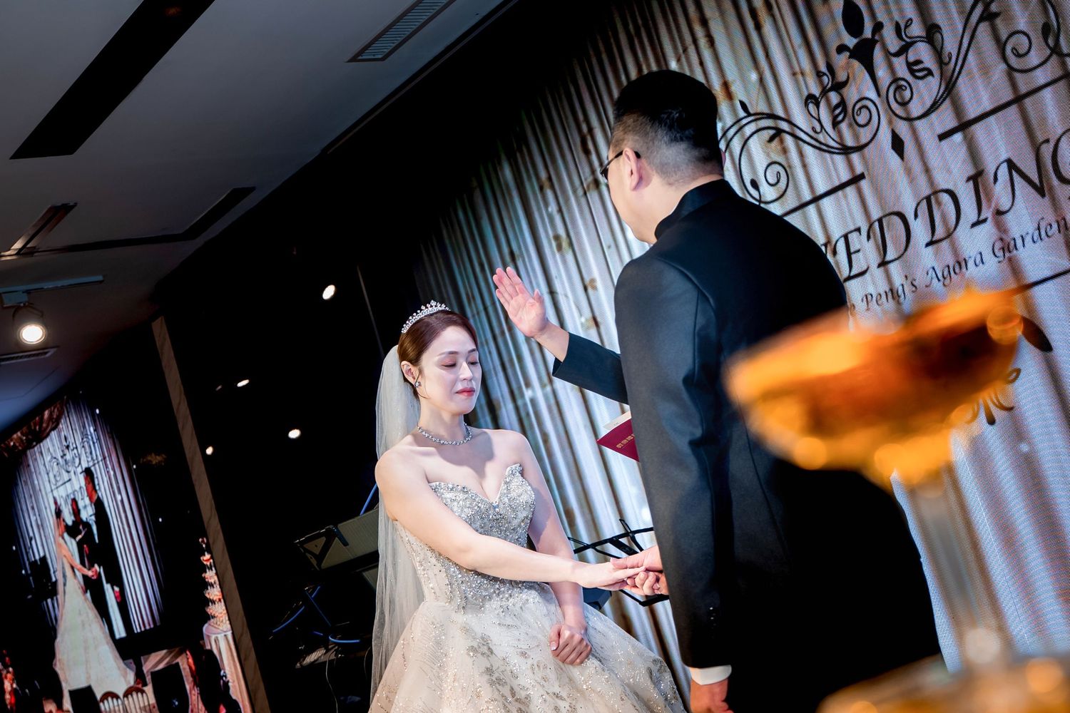 A couple exchanges rings during an elegant indoor wedding ceremony with decorative backdrop and soft lighting.