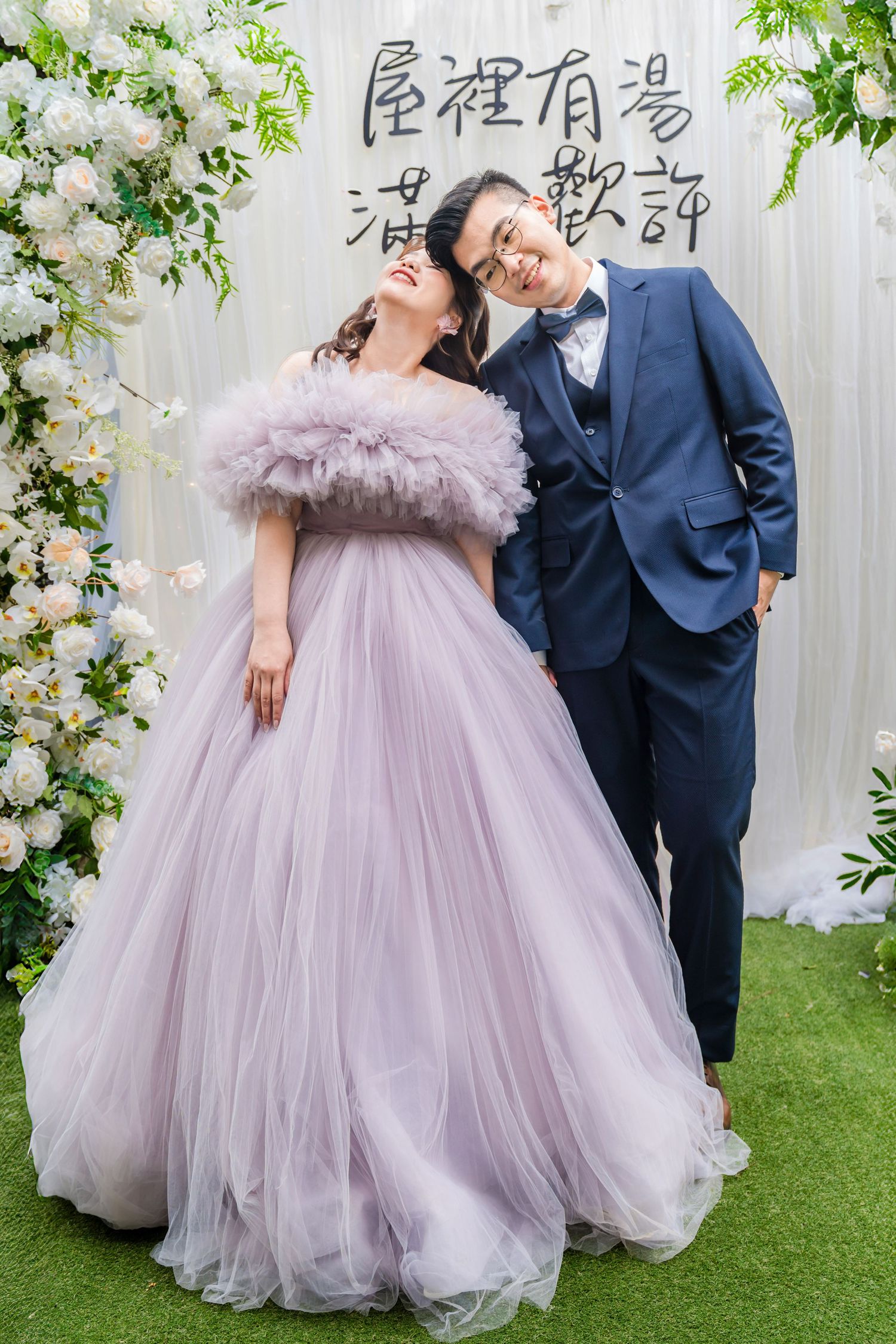 A couple poses in wedding attire - a lavender tulle ball gown and navy suit - against a floral backdrop.