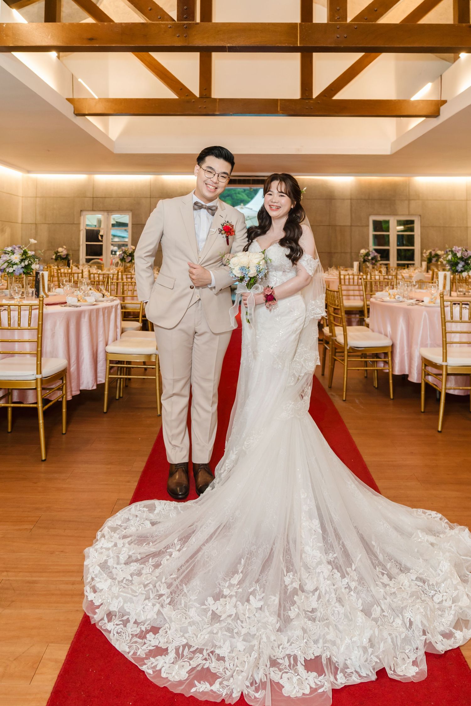 Elegant wedding reception venue with exposed wooden beams, pink linens, gold chairs, and a red carpet aisle.