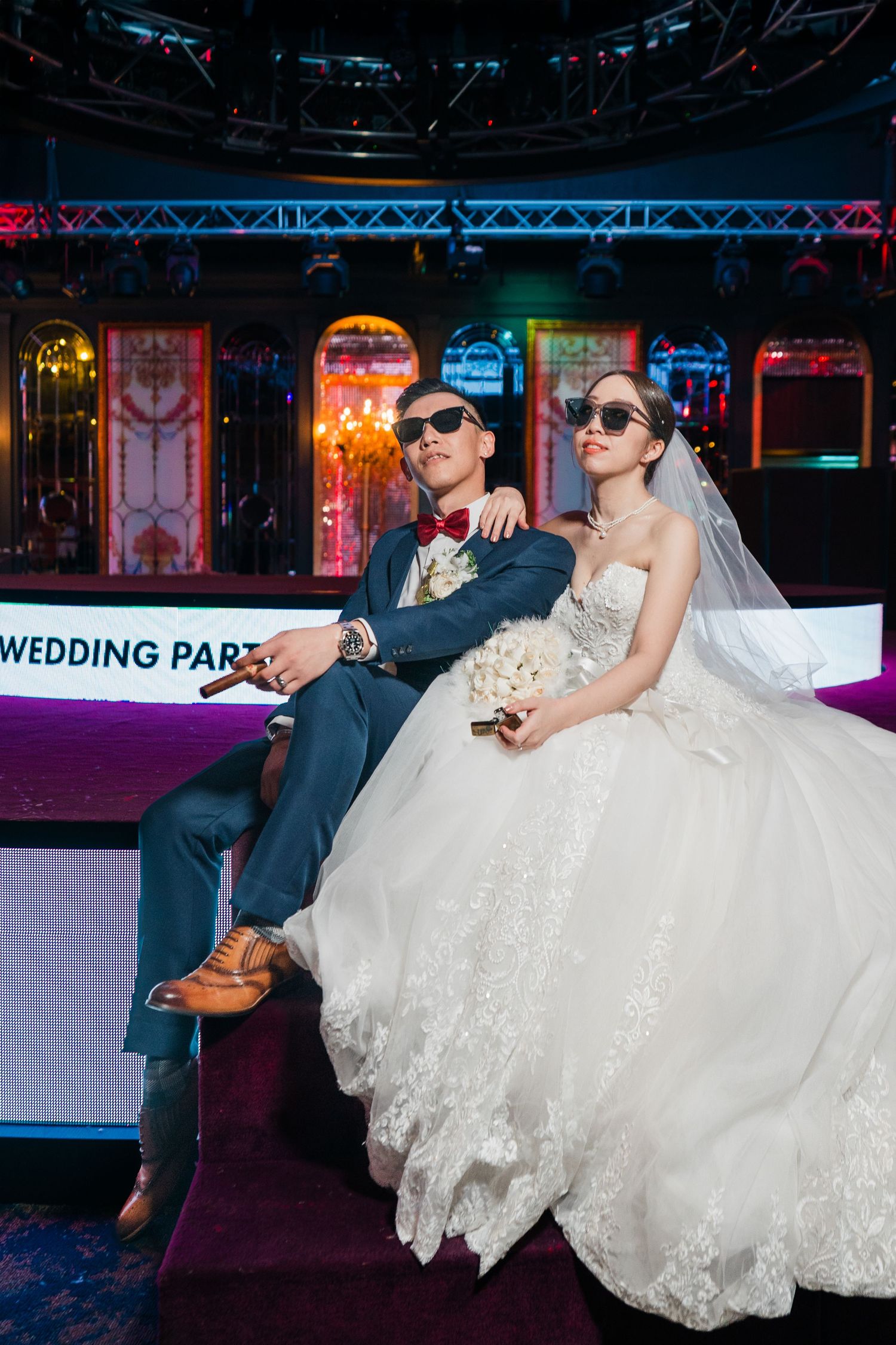 A bride and groom wearing sunglasses relax on a purple couch with neon lights glowing in the background.