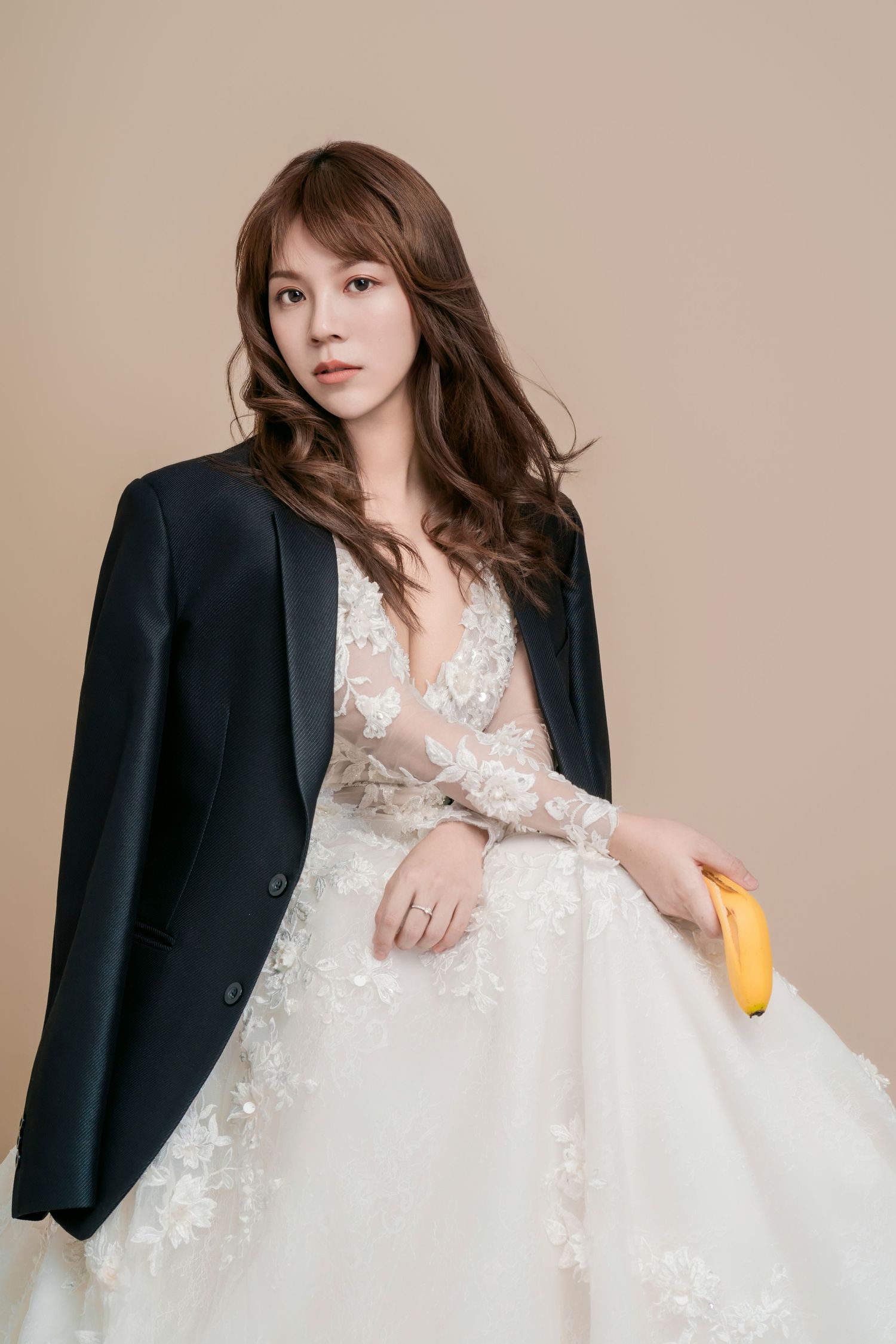 A model in a white lace wedding dress paired with a black blazer poses elegantly against a neutral beige backdrop.
