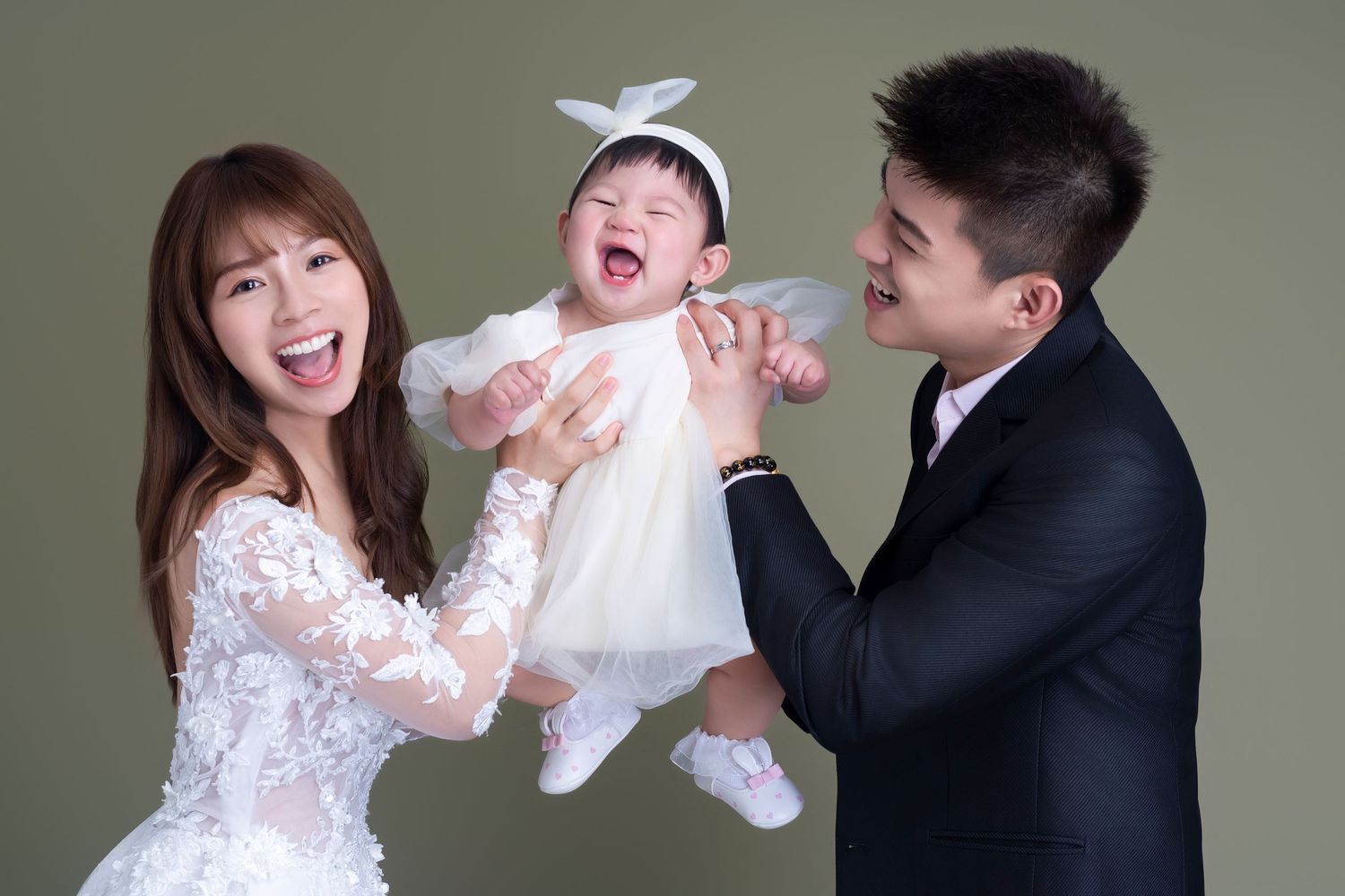 A family in formal attire shares a joyful moment together while holding a laughing baby in a white dress.