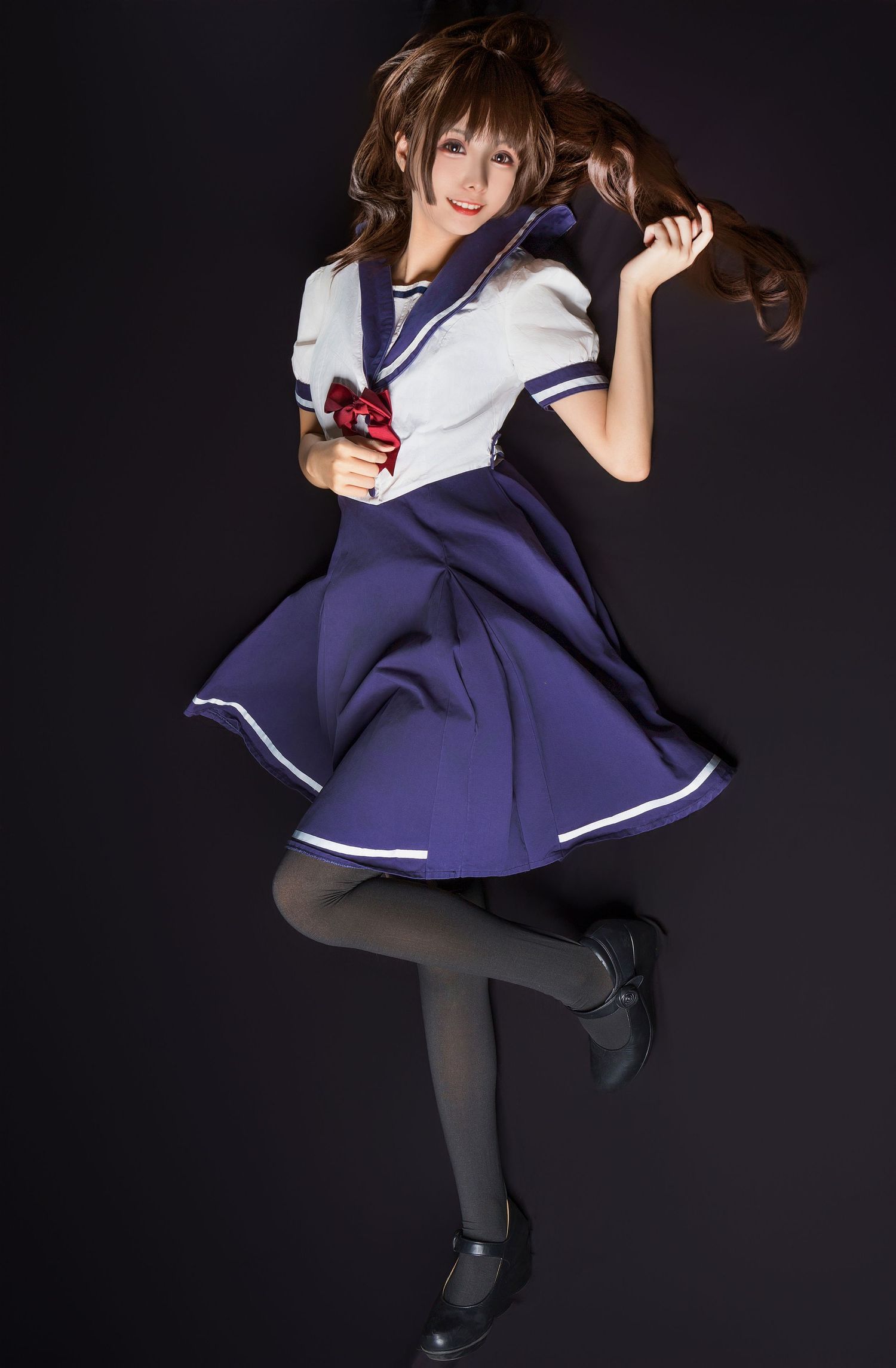 A cosplay model in a navy blue and white sailor-style school uniform dress poses dynamically against a dark background.