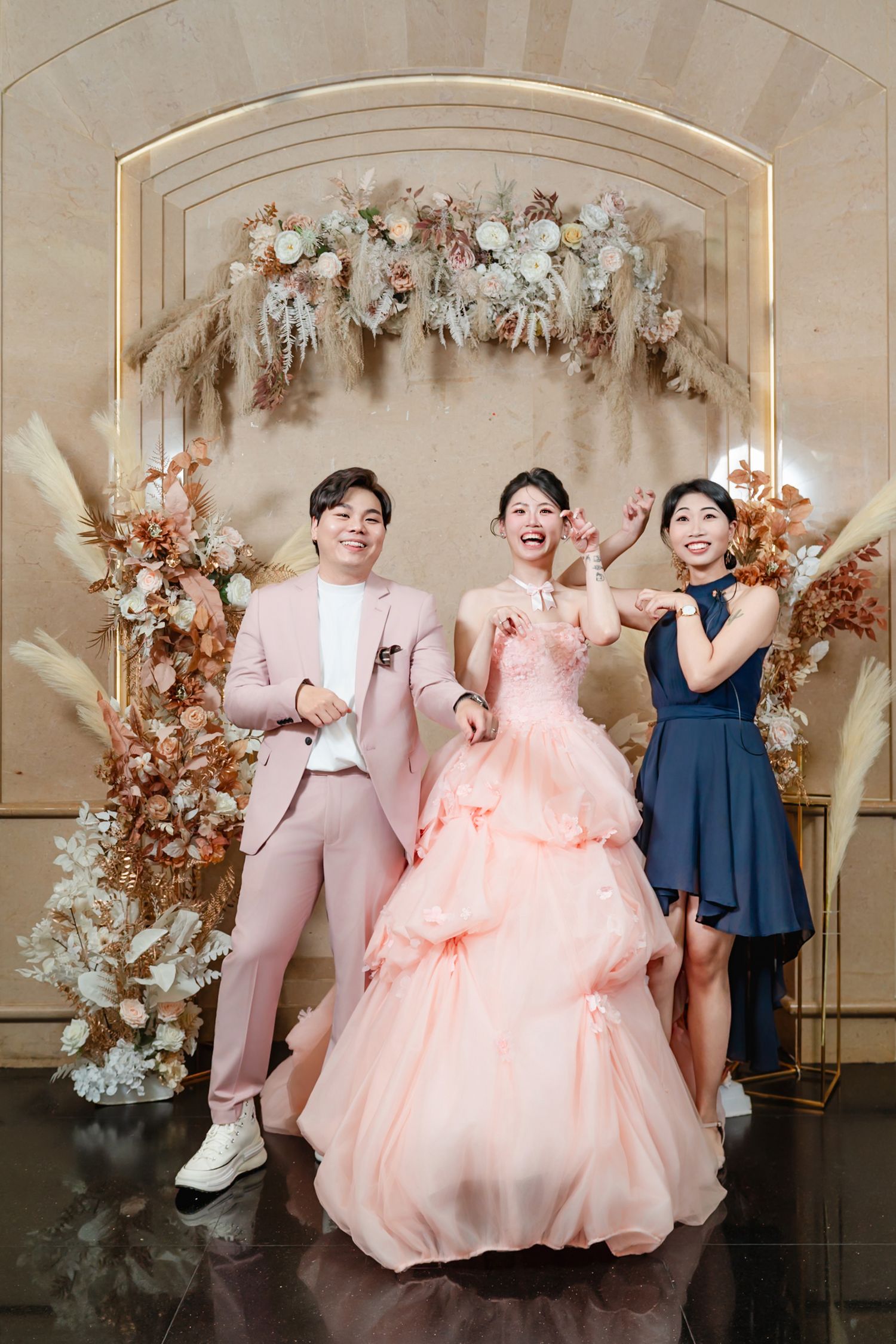 A wedding celebration under an elegant floral arch with pink and white blooms, featuring a peach ballgown and formal attire.