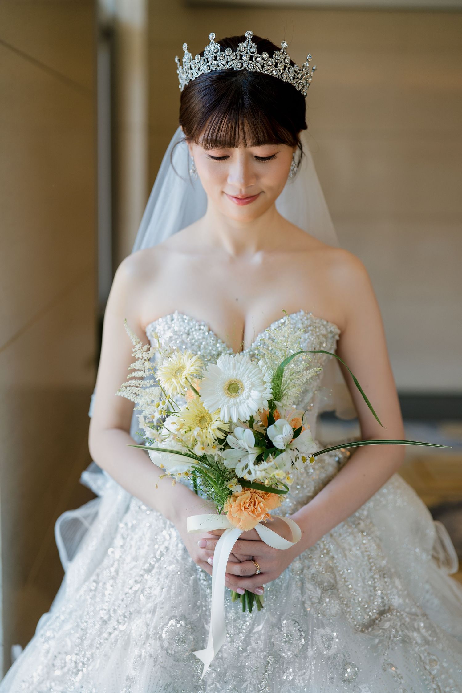 Bride in strapless lace gown and crystal tiara holds yellow and white floral bouquet with ribbon.