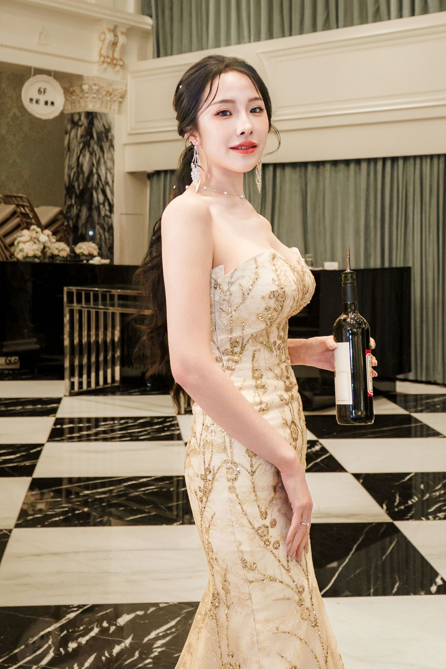 A person poses in an elegant gold sequined evening gown against a luxurious marble and white interior backdrop.