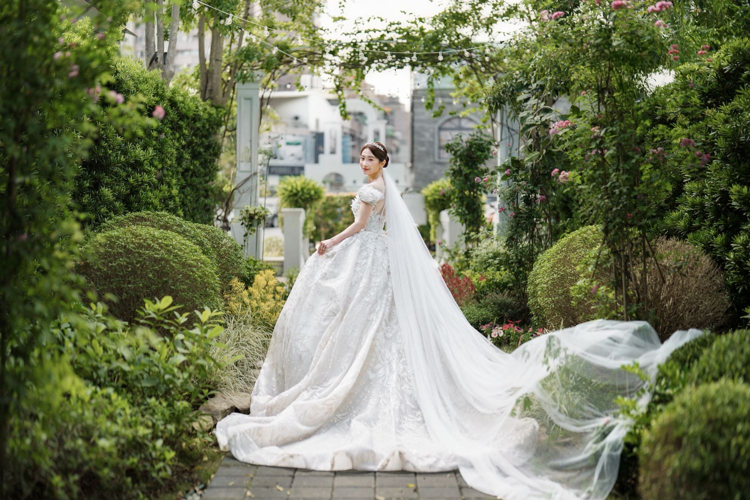 A flowing white wedding gown with a dramatic train captured in a lush garden setting with greenery and stone path.