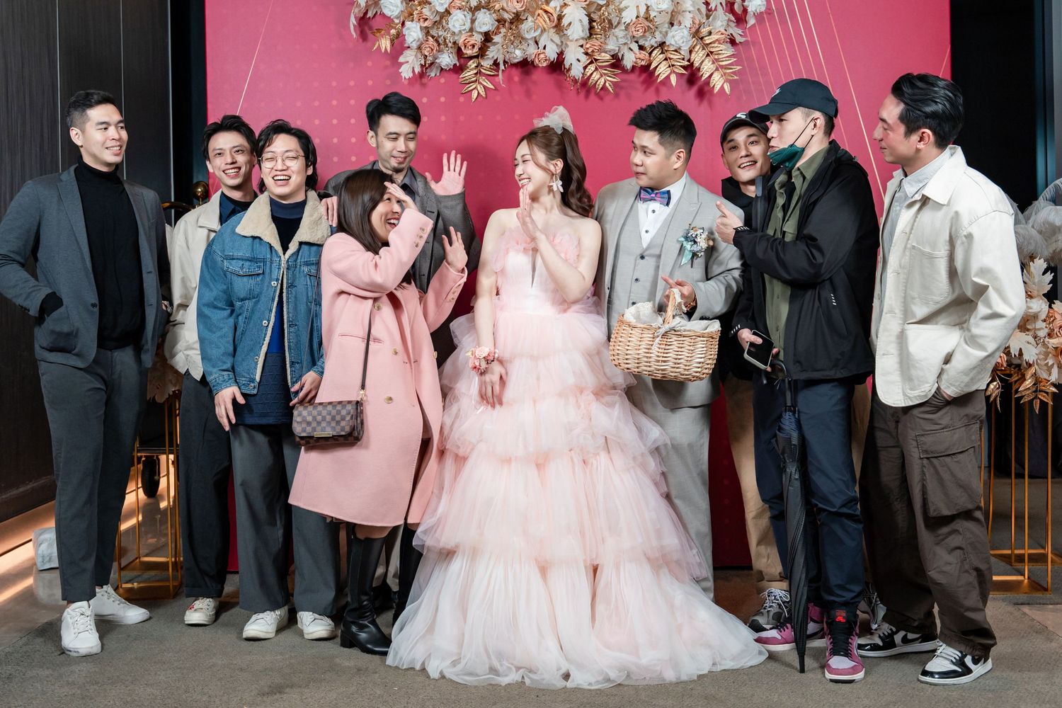 A group of friends pose together at a wedding celebration with a bride in a pink tulle gown under floral decorations.