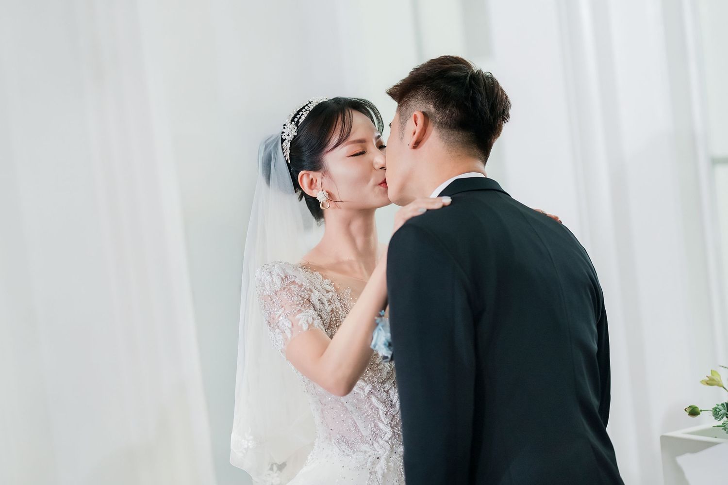 A bride and groom share a romantic kiss during their wedding photoshoot against a white curtain backdrop.
