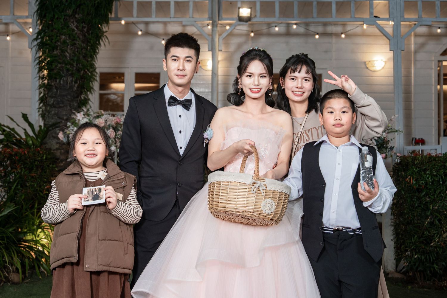 Wedding party guests gather outdoors at evening celebration with decorative lighting and greenery.