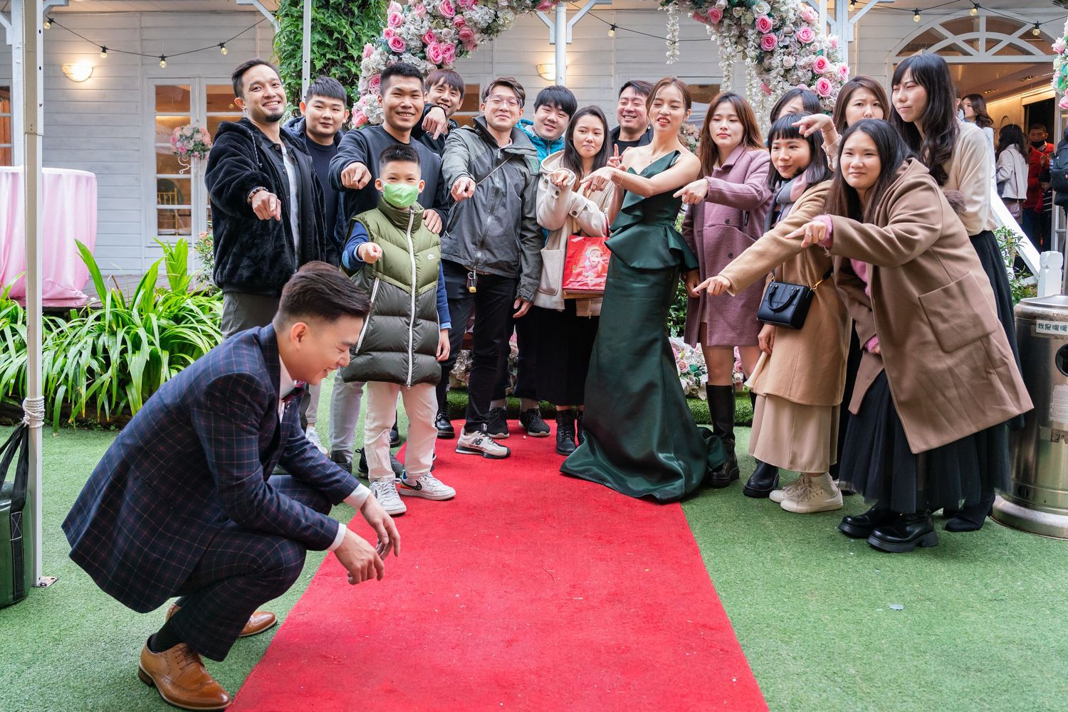 A photographer crouches to take a photo on a red carpet while guests watch and smile at an outdoor event.