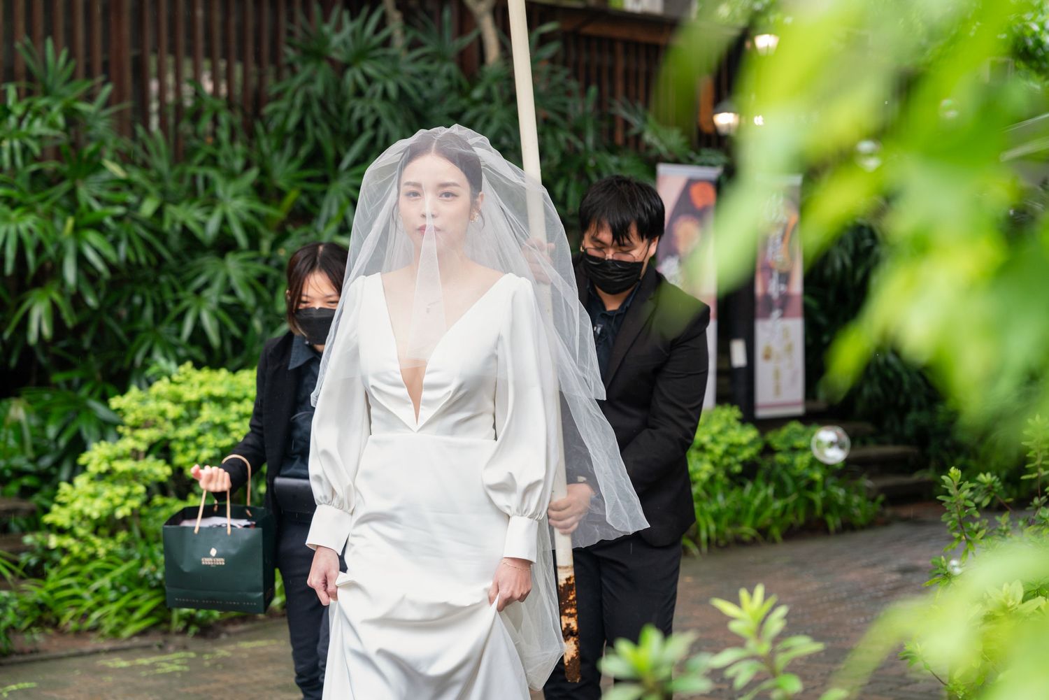 A bride in an elegant white wedding dress and veil stands in a garden setting while others gather behind her.