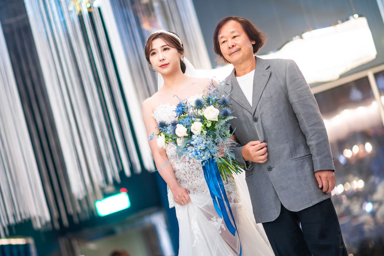Two people in formal attire walking arm in arm while one holds a bouquet with blue accents and ribbons.