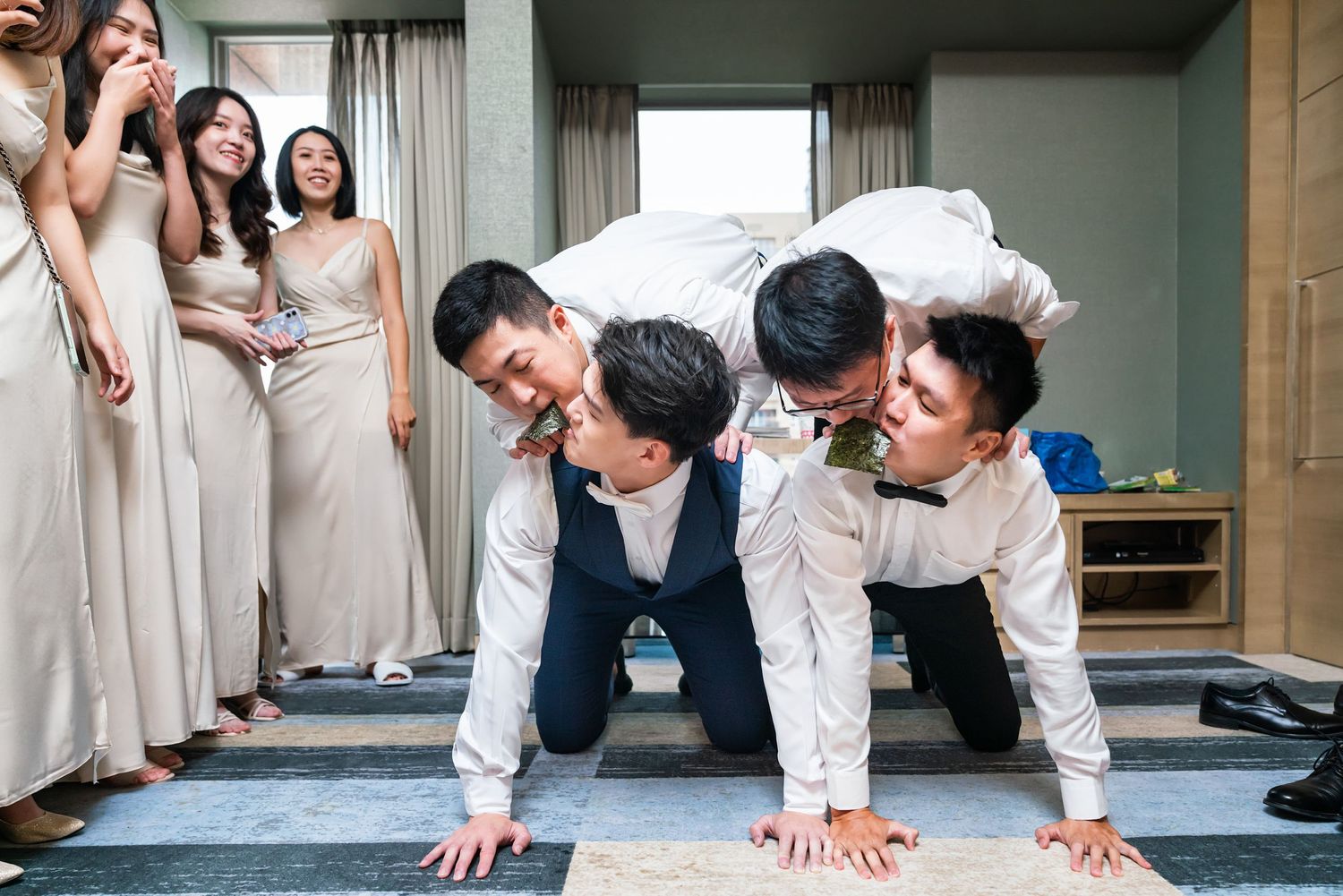 Groomsmen do playful pushups on the floor while bridesmaids in beige dresses laugh and watch from the side.