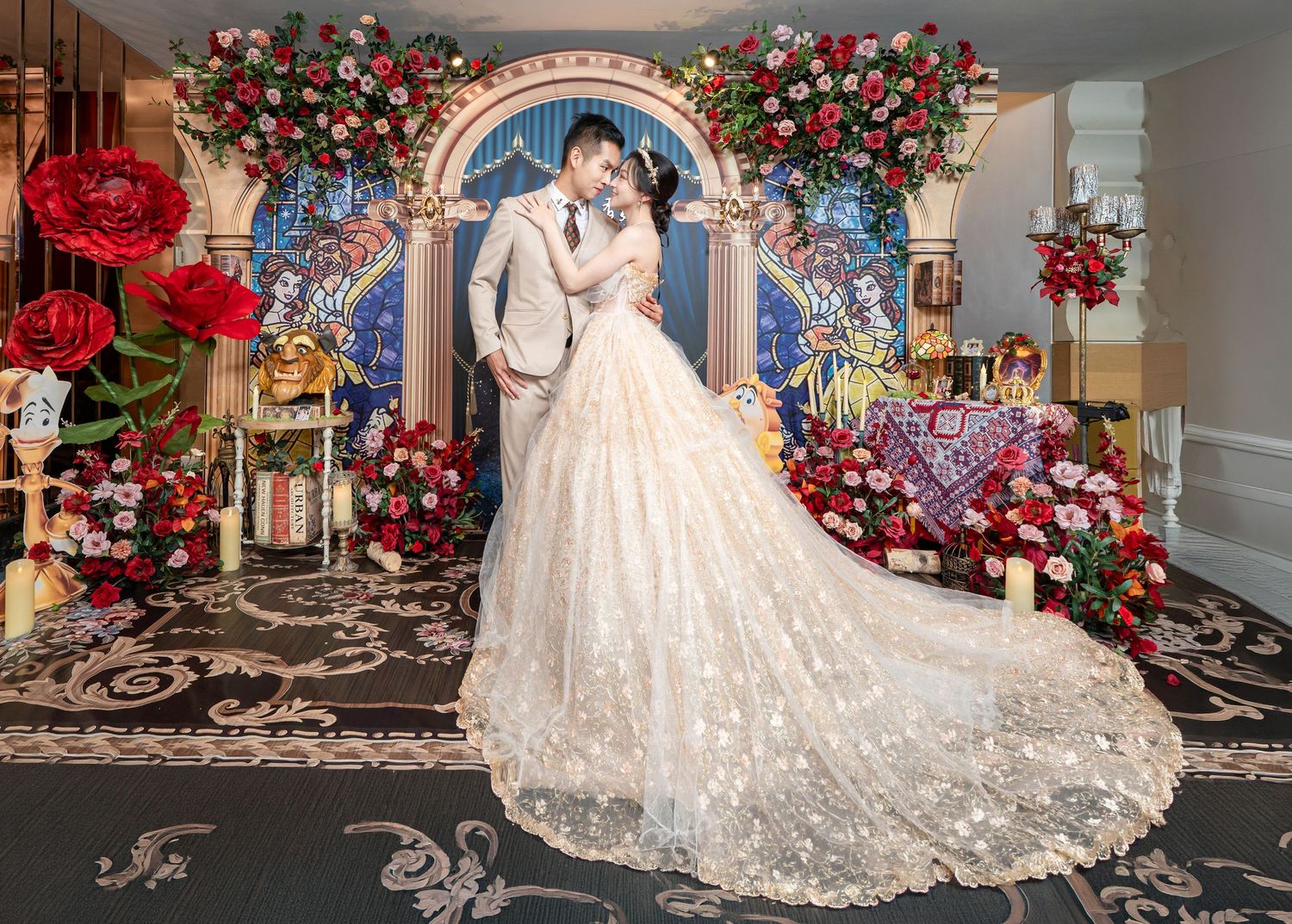 Couple in wedding attire poses before floral arch with red roses and stained glass backdrop in elegant venue.