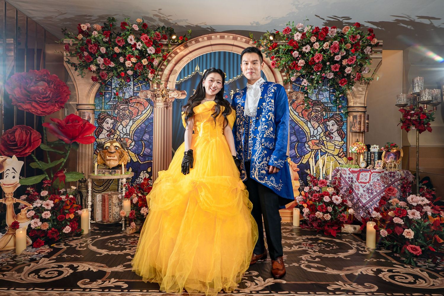 Couple in Beauty and the Beast costumes posing before floral arch backdrop at themed party.