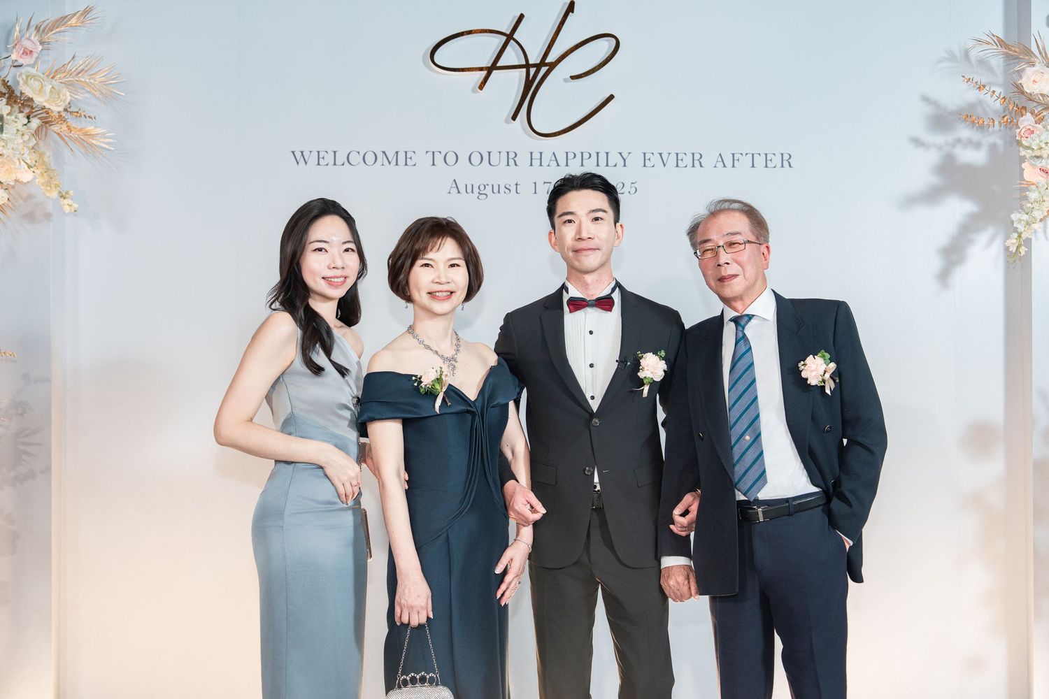 Four people in formal attire pose elegantly at a wedding reception with floral decorations and an elegant monogram backdrop.