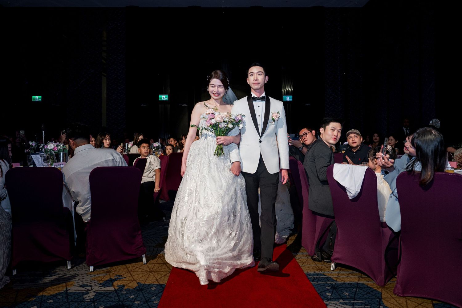 A couple walks down a red carpet at their wedding reception in a dimly lit banquet hall with purple chairs.