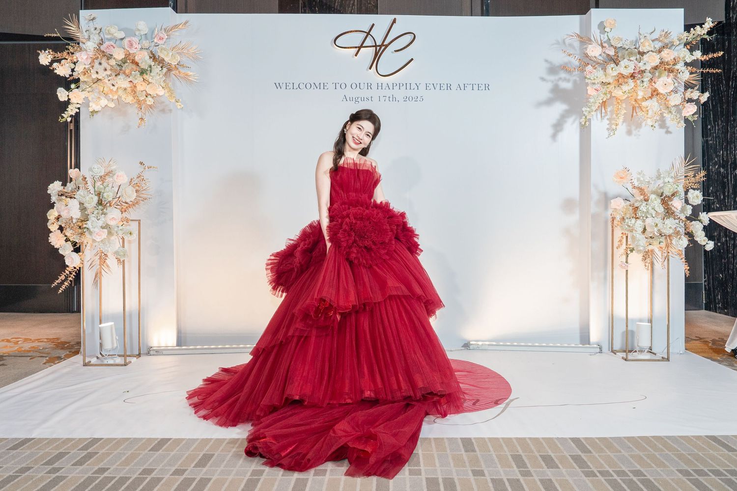 Elegant red tiered ballgown photographed in front of a white backdrop with decorative floral arrangements.