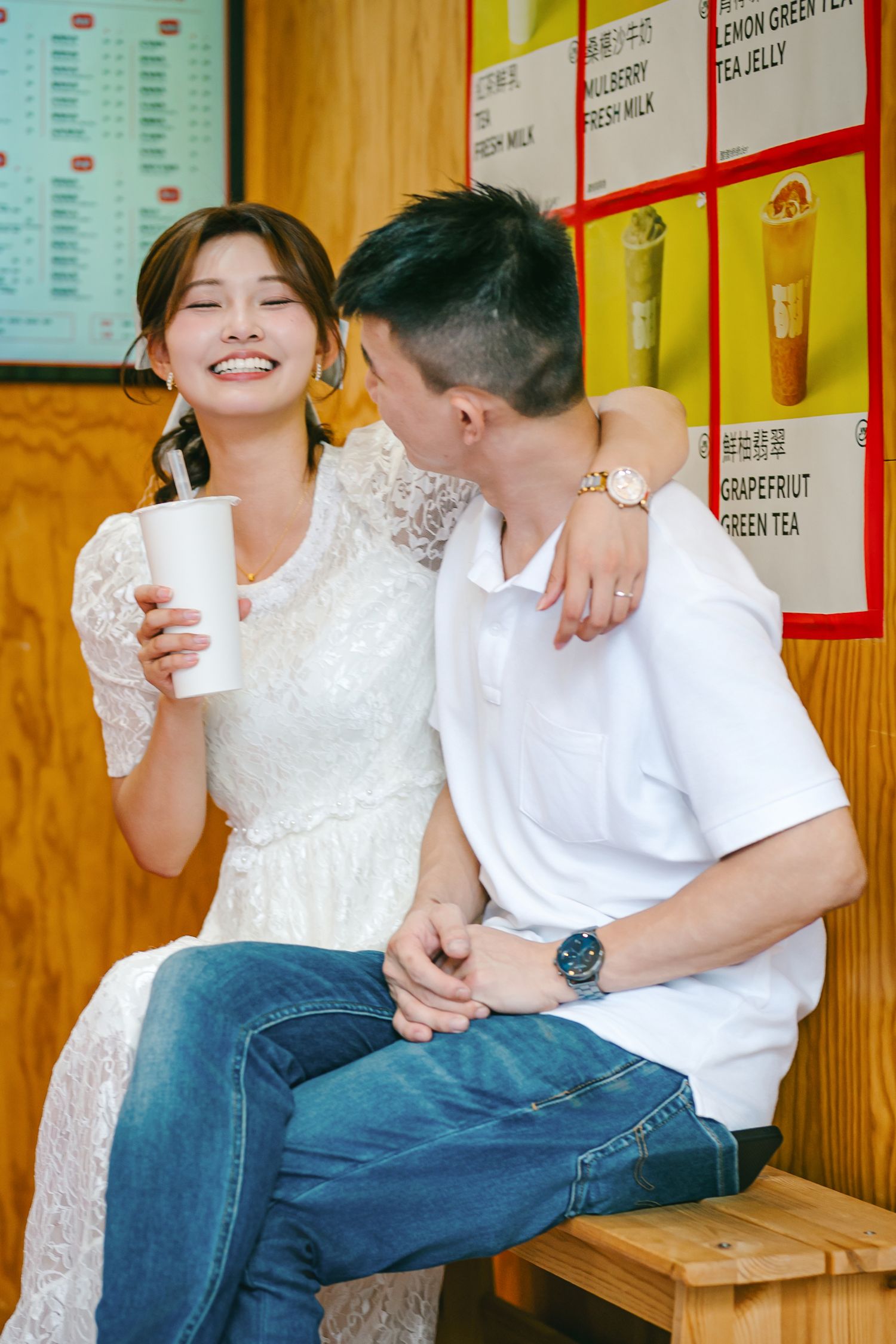 A couple in white tops and jeans sit close together on wooden benches against a wall with colorful menu boards.