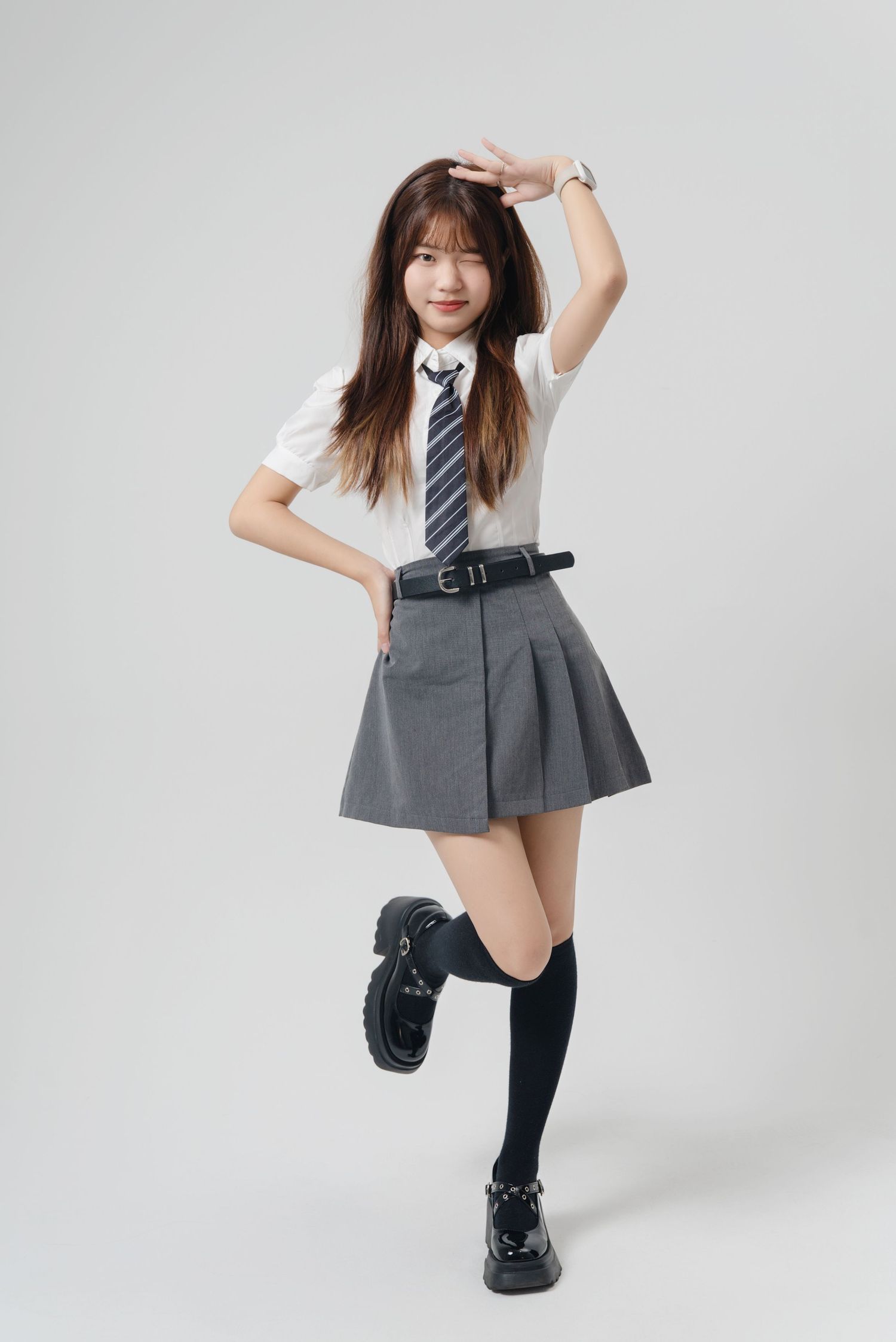 A person in a school uniform with white shirt, gray pleated skirt, tie and knee-high socks poses playfully against white background.