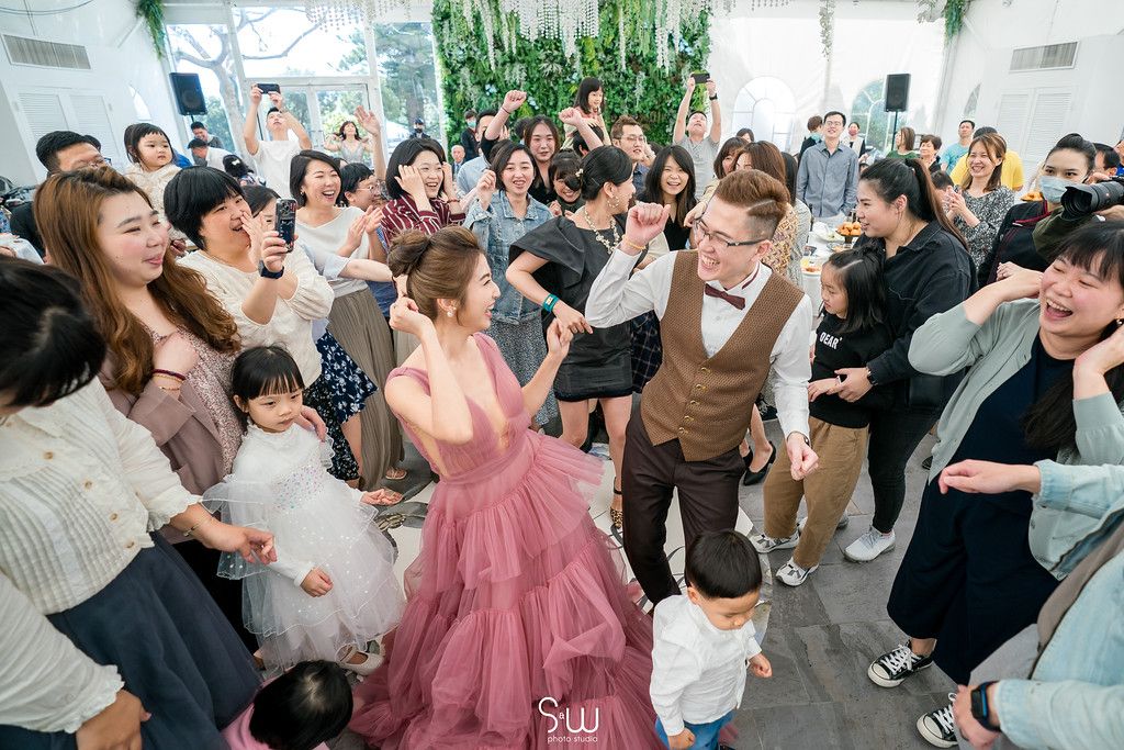A joyous celebration in a light-filled venue with hanging greenery as guests dance and laugh together.