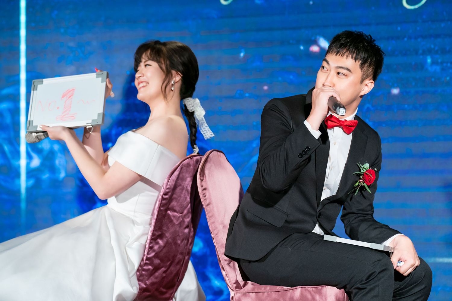 A couple in wedding attire pose back-to-back on stage during a festive celebration.