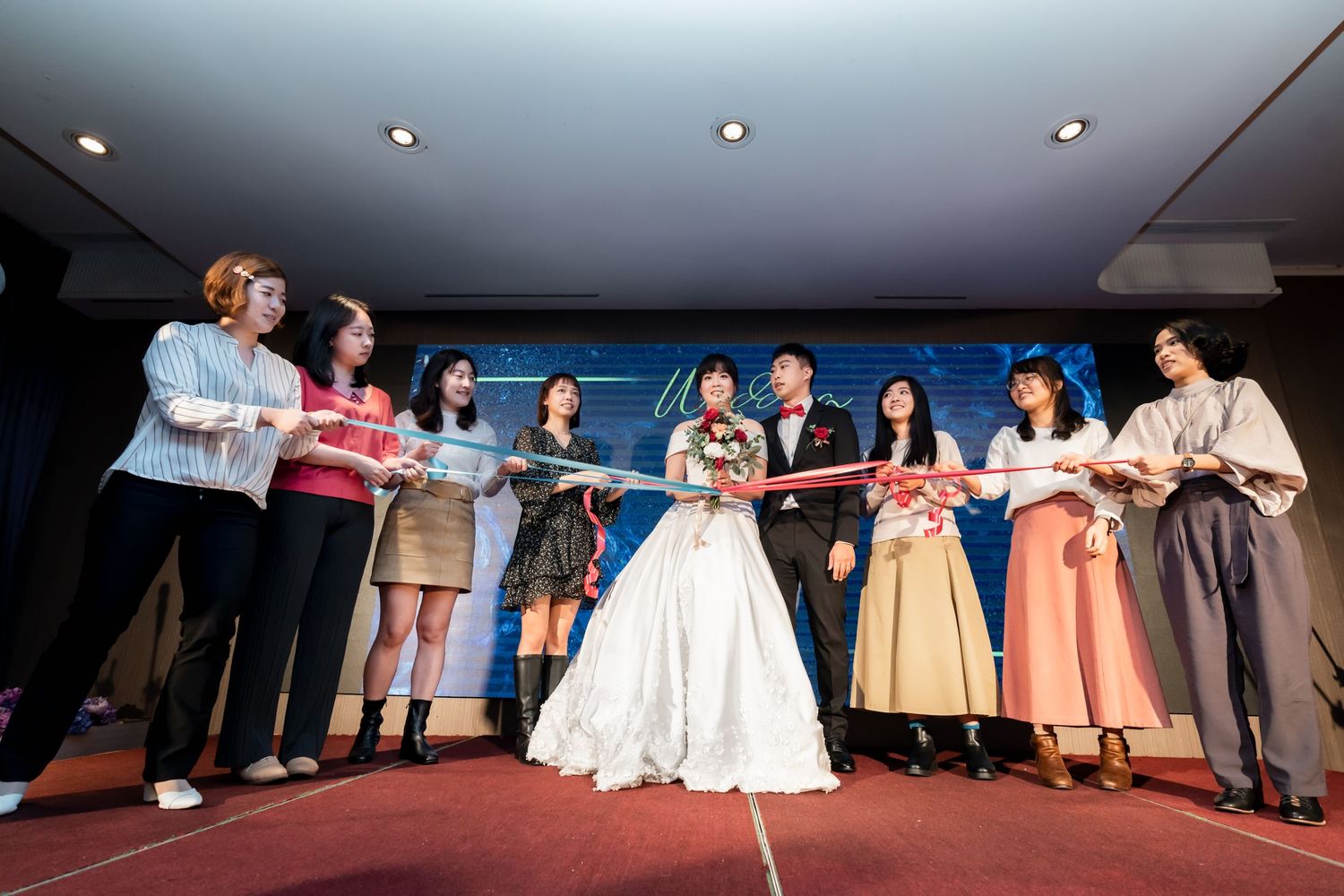 Wedding party lines up on stage with bride in white gown at center.