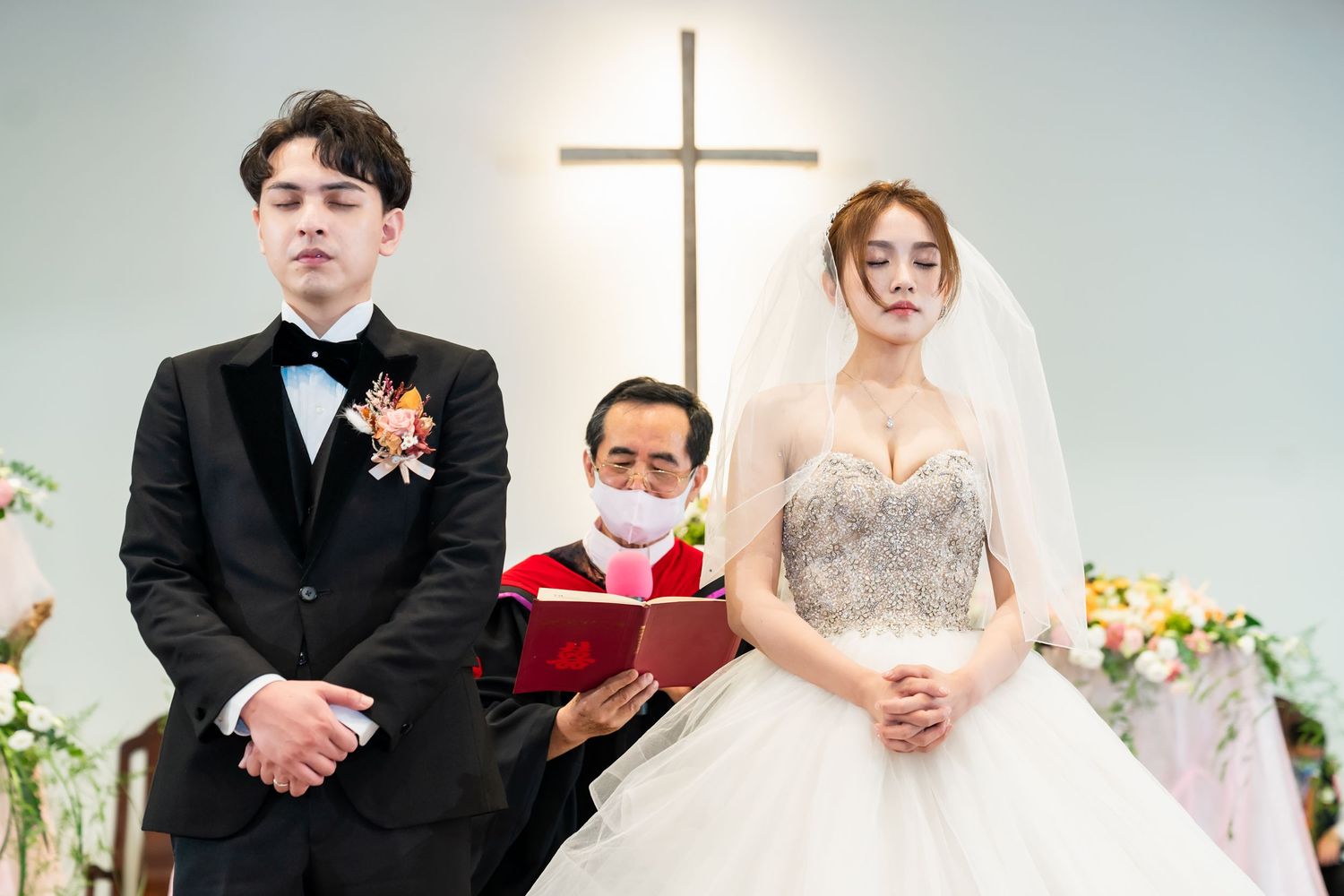 A wedding portrait of a couple and officiant standing beneath a cross in a church.