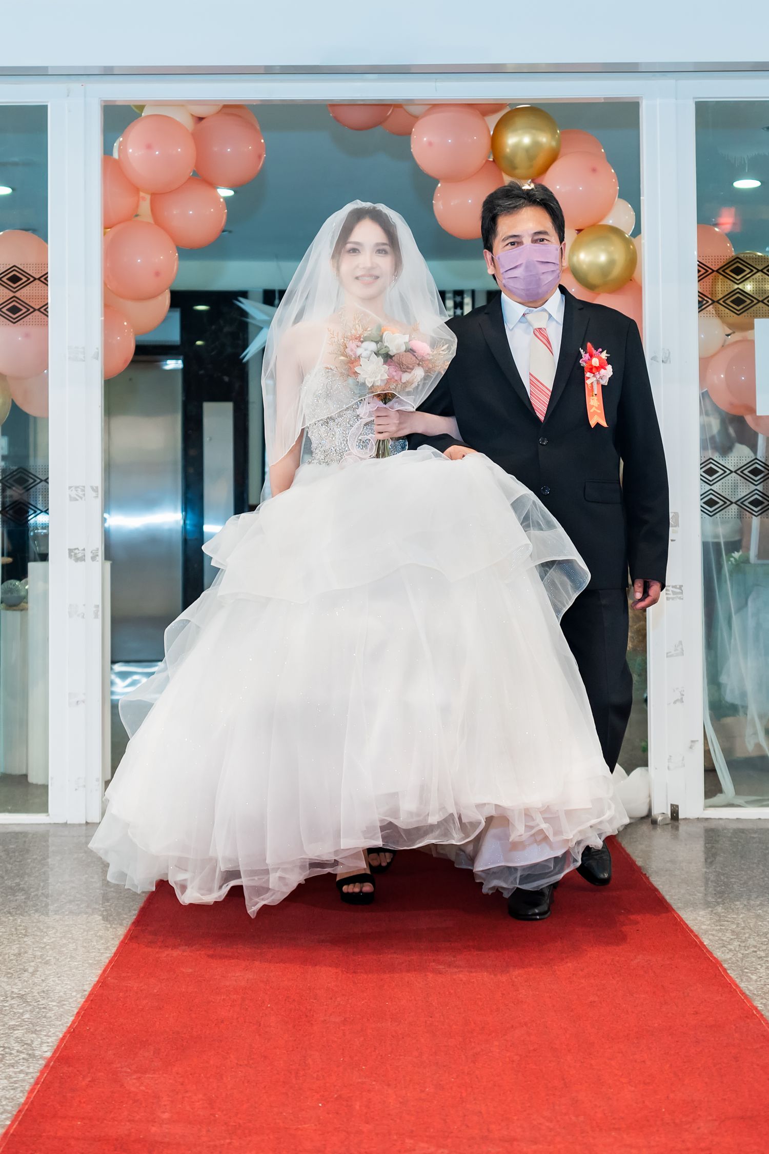 A couple in wedding attire pose on red carpet under coral and gold balloon decorations.