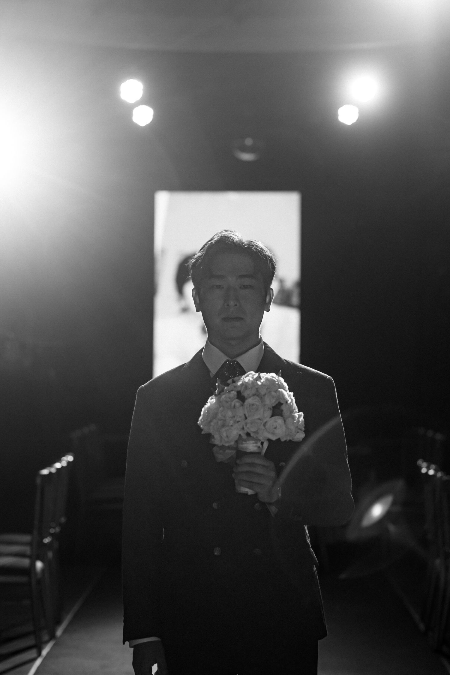Black and white photo of a man in a suit holding a bouquet of flowers in a dimly lit venue.
